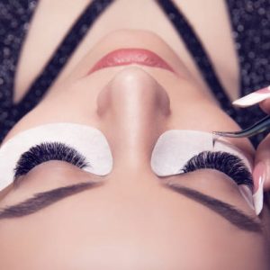 Eyelash extension process. Closeup portrait of young girl, woman with long and thick eyelashes, eyes closed and hand of a cosmetologist adding more eyelashes to her.